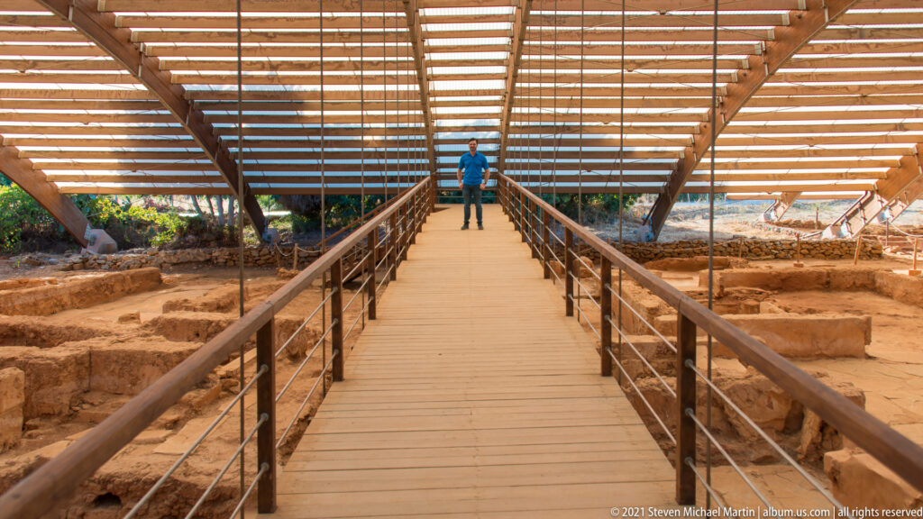 Wooden visitor walkway with railings beneath a slatted shelter roof, flanked by excavated ruins.