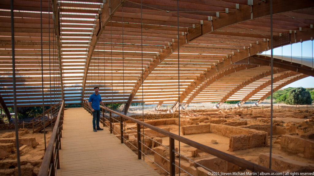 Visitor on a raised walkway beneath a large slatted shelter roof above excavated palace remains.