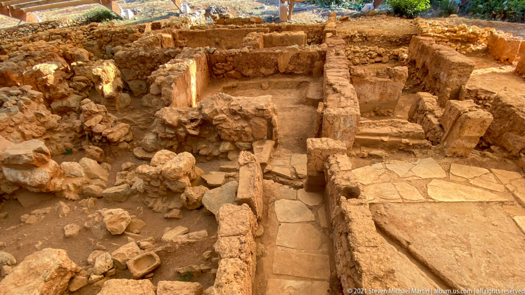 Narrow stone-paved passage between low excavated walls and adjoining rooms at Malia Minoan Palace.