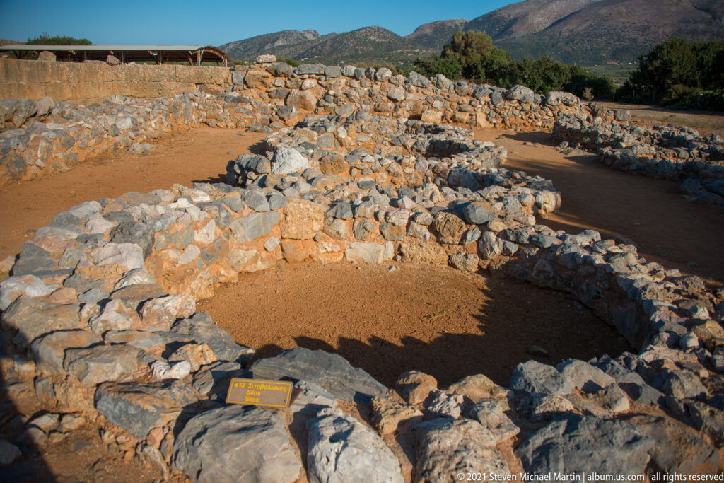 Low circular stone enclosure in the foreground with excavated walls and mountains behind.