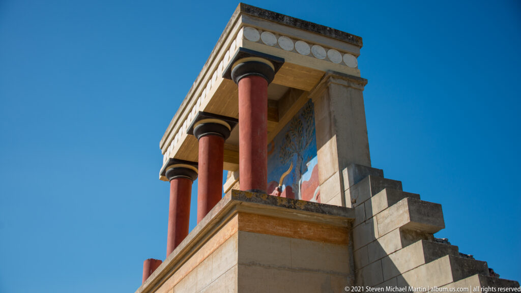 Low-angle view of the reconstructed North Entrance at Knossos with red columns, pale entablature, and a bull fresco panel.