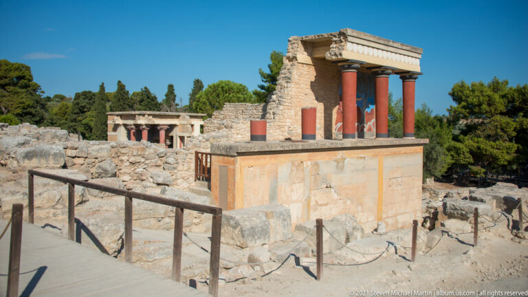 North Entrance reconstruction with red columns and bull fresco amid extensive Knossos ruins, viewed from a visitor walkway.