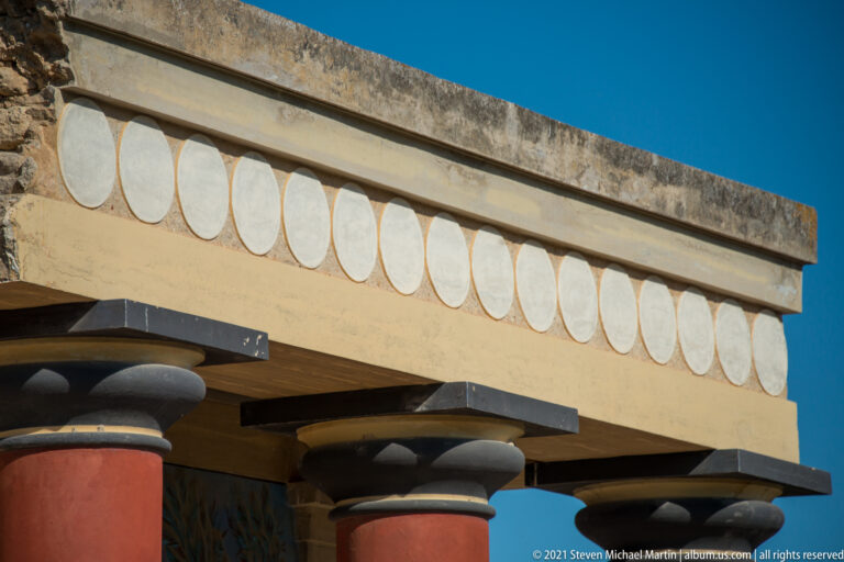 Close view of circular disc motifs on the North Entrance entablature above dark capitals and red column tops.
