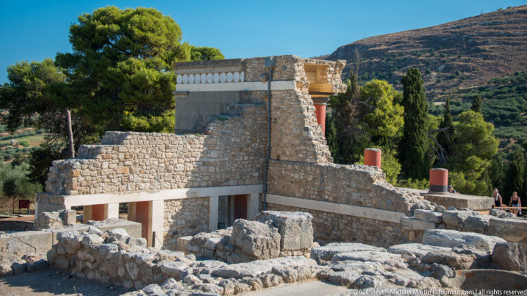 Reconstructed North Entrance area rising above excavated stone foundations, with red column elements and a hillside beyond.