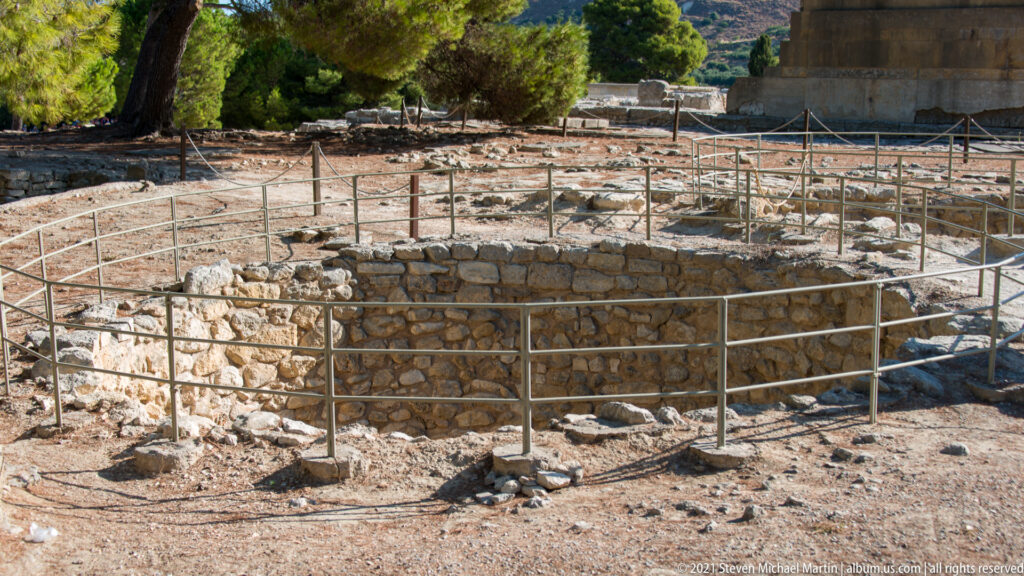 Circular stone-lined kouloura at Knossos surrounded by protective railings and low archaeological masonry.