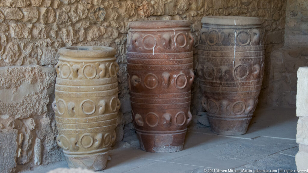 Three tall Minoan-style pithoi with raised bands and looped relief decoration standing against a rough stone wall.