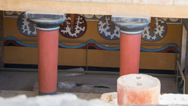 Repeated figure-eight shield motifs painted on an interior wall behind two reconstructed red Minoan columns.