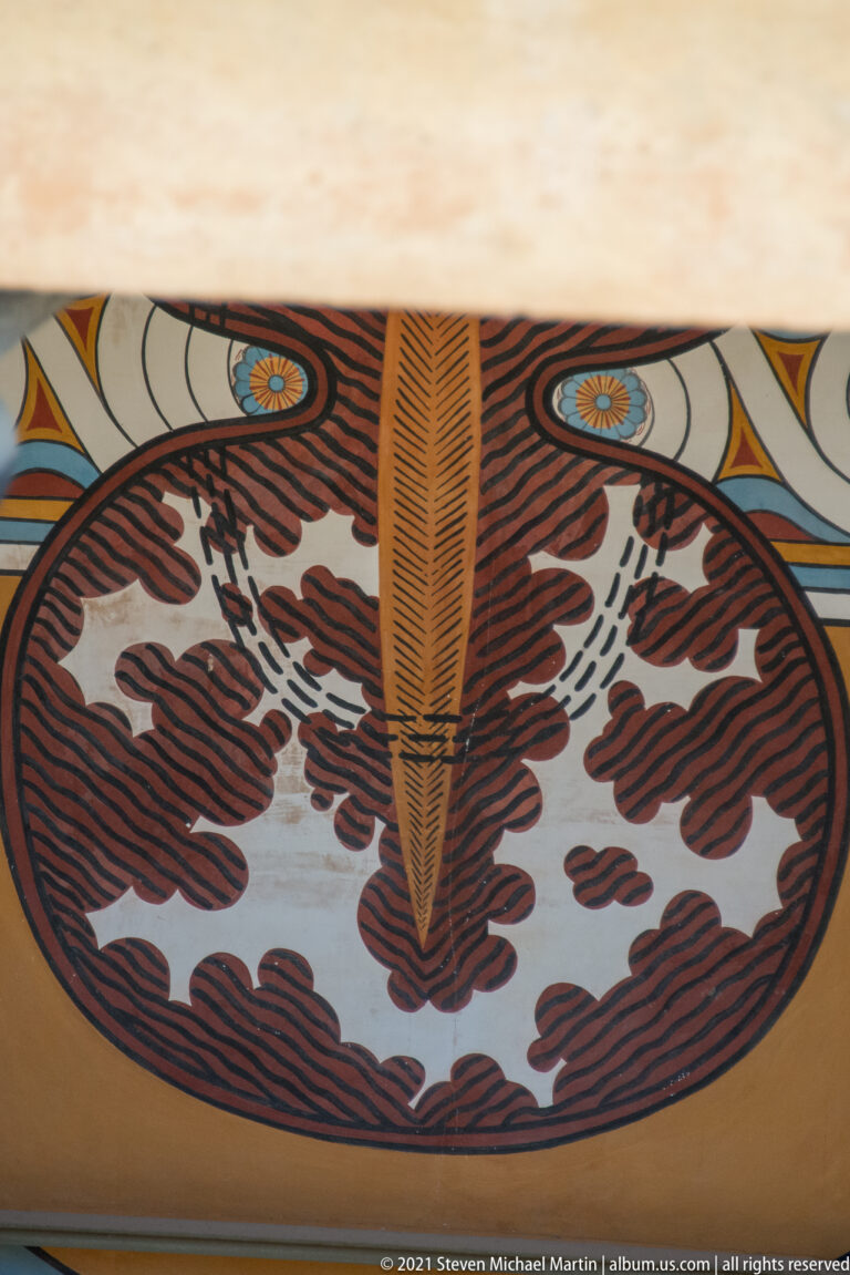 Close detail of a painted figure-eight shield motif with dark red, black, cream, and blue decoration.