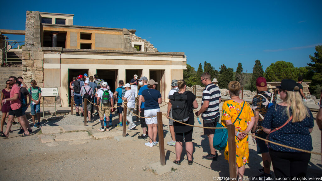 Visitors waiting in a roped queue beside reconstructed palace walls and excavated stonework at Knossos.