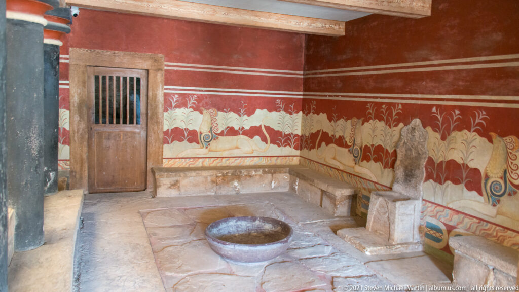 Throne Room at Knossos with griffin frescoes, a stone seat, red walls, and a circular stone basin in the floor.