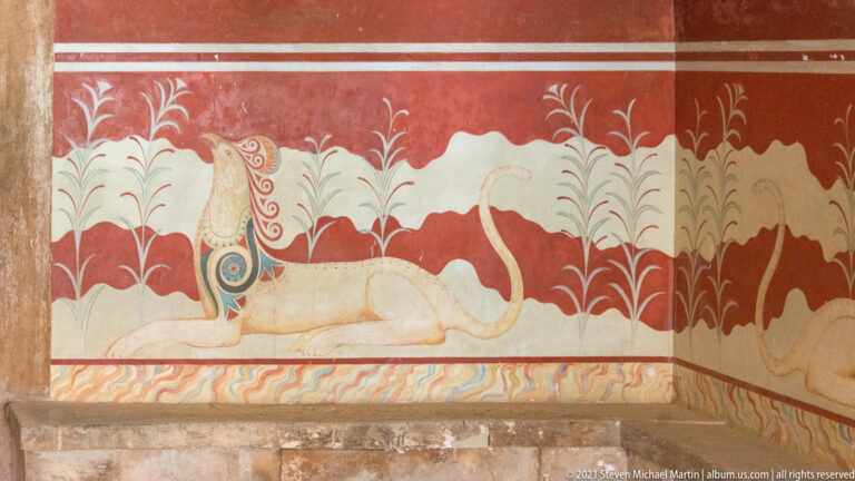 Reclining pale griffin with a patterned wing and curled tail among stylized plants on the red Throne Room wall.