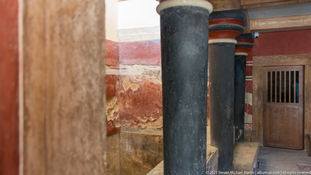 Black-painted columns with colored capitals beside restored red walls, exposed stonework, and a barred wooden door.