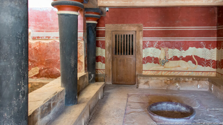 Red reconstructed room with black columns, griffin frieze, barred door, benches, and a round floor basin.