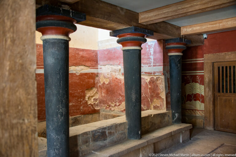 Three black columns with colored capitals beside red restored walls, stone benching, and a wooden door.
