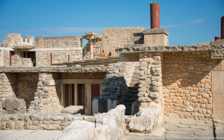 Excavated stone walls surrounding a lower reconstructed doorway and columns, with a tall red column on the upper level.
