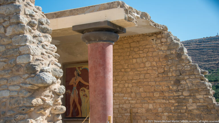 Red tapered column supporting a reconstructed lintel beside rough stone walls, with a fresco figure visible behind.