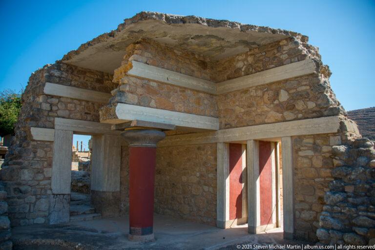 Reconstructed stone palace corner with pale beams, a red column, and open doorways at ground level.