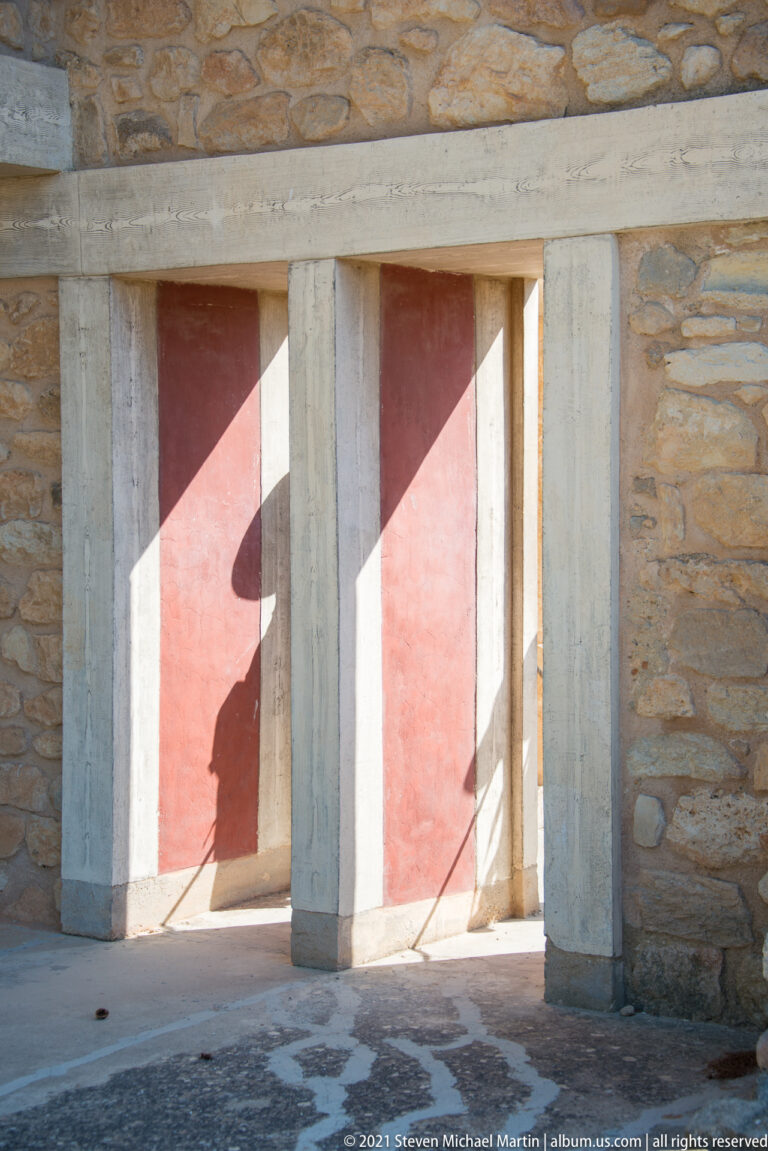 Three pale vertical piers forming a broad opening in a stone wall with red recessed panels and sharp shadows.