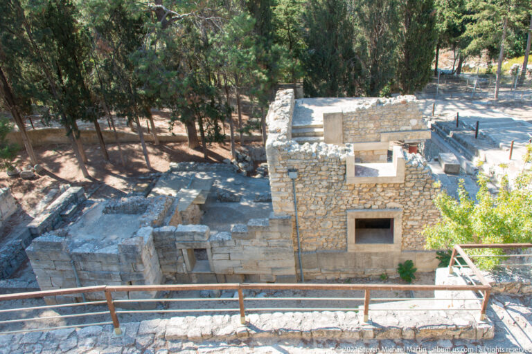 Elevated view over excavated stone rooms, steps, walls, railings, and shaded trees at Knossos.