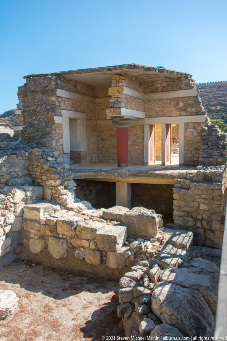 Low stone façade with pale bands, a red column, deep openings, and excavated wall foundations in front.