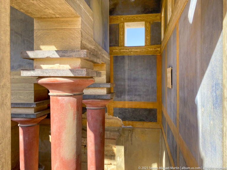 Several red tapered columns beneath heavy stepped beams, with a small high window opening to blue sky.