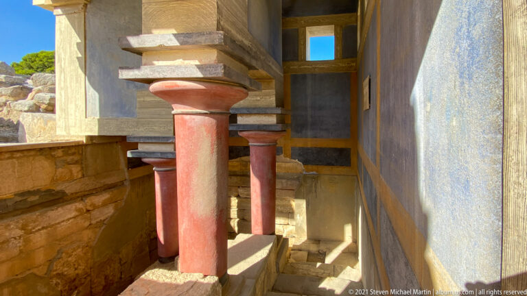 Red columns beside descending steps and heavy beams in a sunlit reconstructed palace interior.