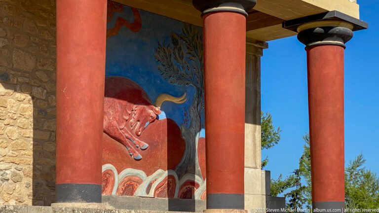 Reconstructed red bull fresco on a blue ground between two red columns, with stylized vegetation behind.
