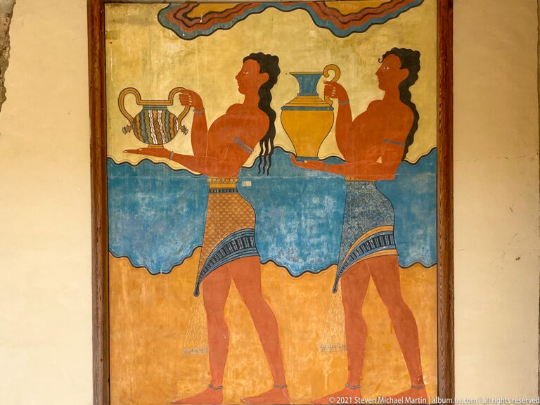 Two reconstructed Minoan procession figures carrying vessels, bordered by red, blue, and black decorative bands.