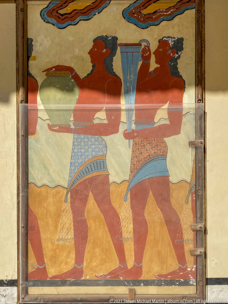 Two profile figures in patterned garments beside large vessels in a reconstructed Minoan procession fresco behind glass.