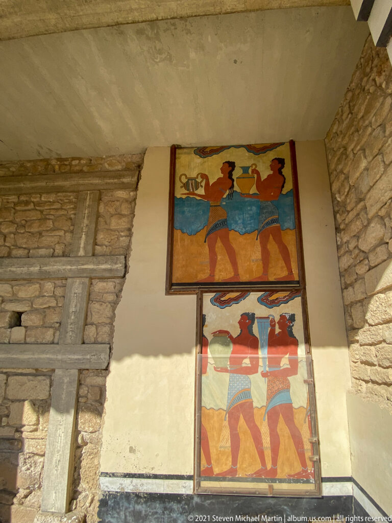 Two stacked procession fresco reconstructions mounted on a pale wall beside exposed stone masonry.