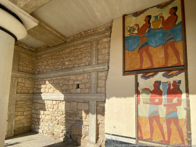 Stacked procession fresco panels beside a broad sunlit wall of rough stone masonry and reconstructed framing.