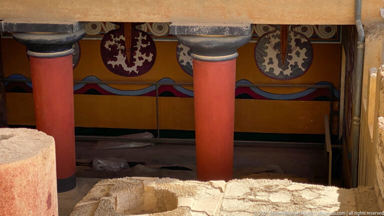 Two red columns in front of painted figure-eight shield motifs, with excavated stone foundations in the foreground.