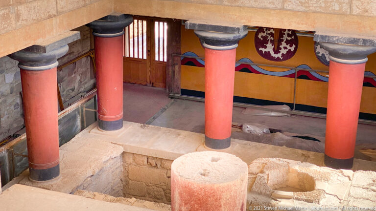 Three red columns before a painted wall with figure-eight shield motifs, doorway, and excavated masonry below.