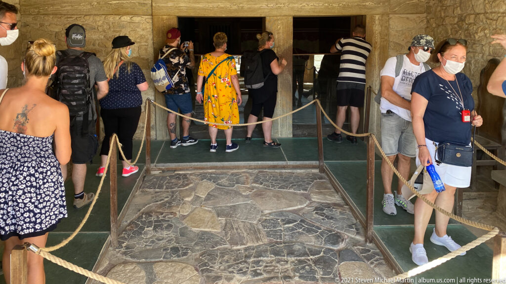 Visitors on roped walkways entering a sheltered archaeological interior over a stone-paved surface at Knossos.