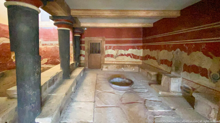 Long red painted chamber with black columns, griffin frieze, stone basin, benches, and a barred door.