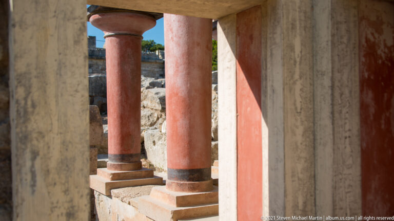 Close view of red tapered Minoan columns beside pale reconstructed walls and rough stone masonry at the North Lustral Basin.