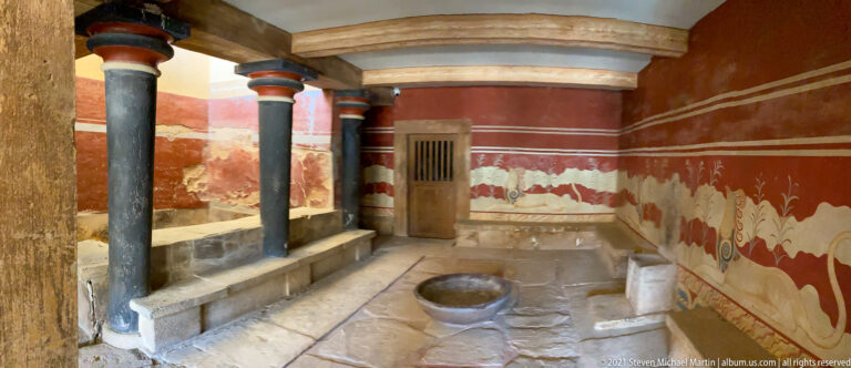 Wide view of a red reconstructed chamber with black columns, griffin decoration, central stone basin, and doorway.