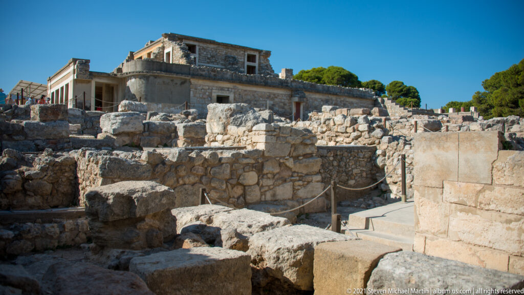 Excavated stone walls and foundations at Knossos with reconstructed palace architecture rising behind them under a clear sky.