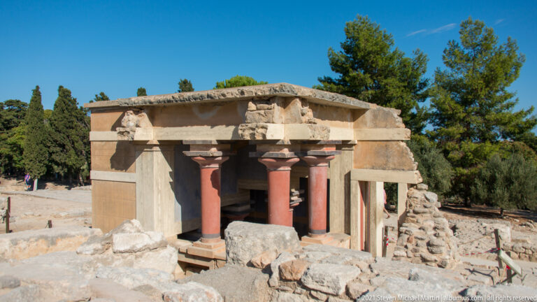 Overall view of the North Lustral Basin reconstruction with paired red columns, pale beams, side walls, and surrounding ruins.