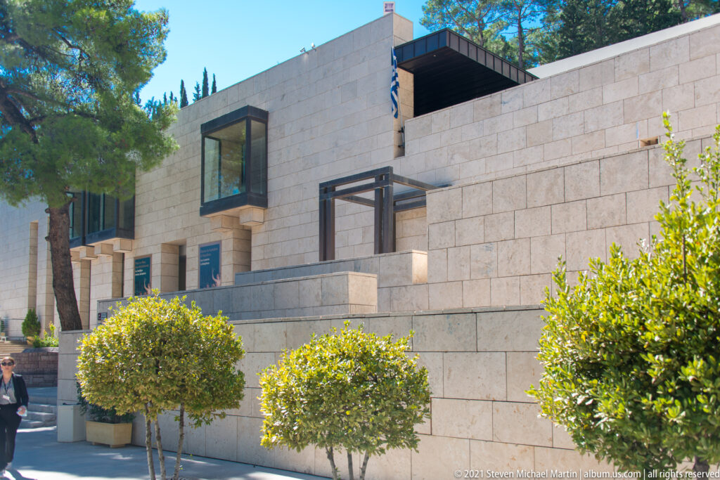 Pale-stone exterior of the Delphi Archaeological Museum with projecting windows, trees, shrubs, and a Greek flag.