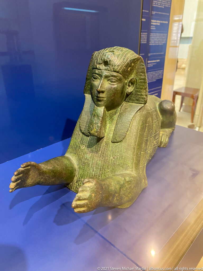 Reclining sphinx with lion body, human head, Egyptian-style headdress, and extended forelegs in a museum case.