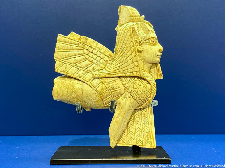Profile view of a small winged sphinx with human head, feline body, and patterned wings on a museum mount.