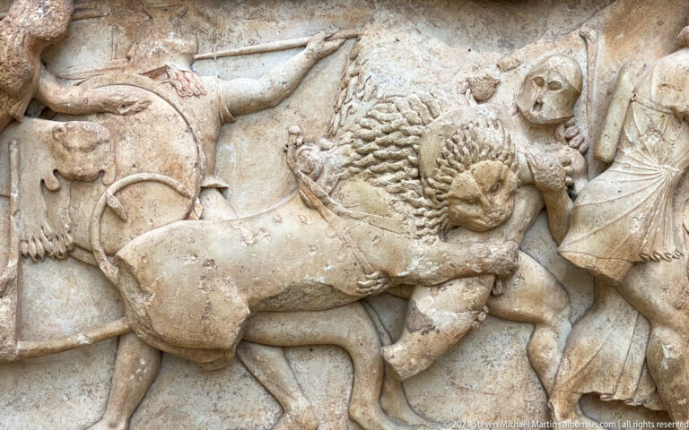 Stone relief with a lion attacking a human figure amid overlapping bodies and surviving fragments of other figures.