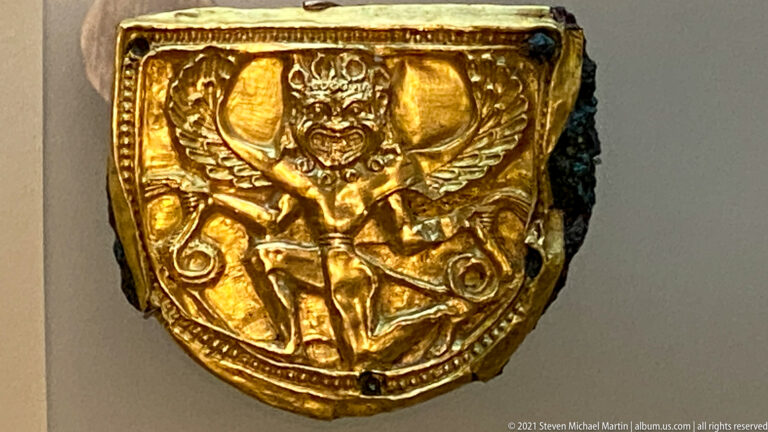 Shield-shaped gold plaque embossed with a frontal winged figure surrounded by scrolling ornament.