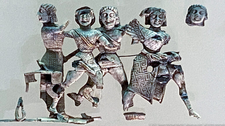 Group of thin metal cutout figures with patterned clothing, separate heads, and overlapping limbs in a museum display.