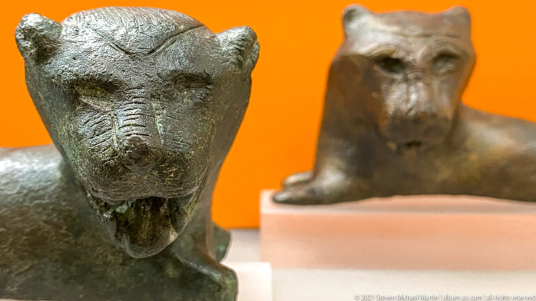 Close view of a bronze lion head with rounded ears and open mouth, with a second feline fitting blurred behind.