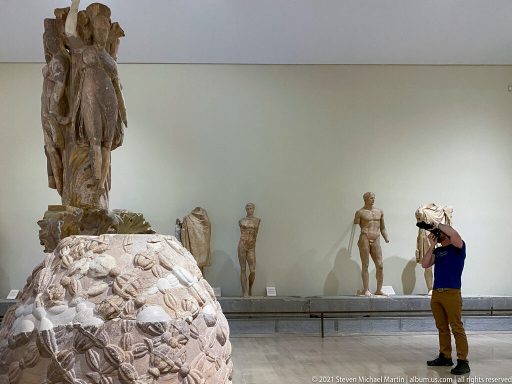Visitor photographing a museum gallery with standing ancient statues and a large carved marble monument in the foreground.