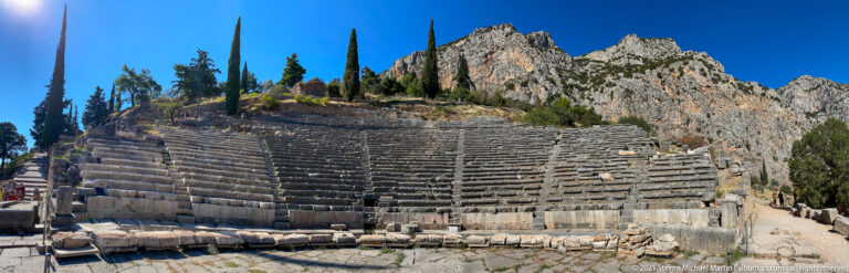 Wide frontal view of the Theatre of Delphi, with curved stone seating, stairways, cypress trees, and rocky peaks.