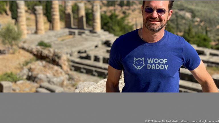 Smiling man in sunglasses and a blue T-shirt standing before blurred stone ruins and columns at Delphi.