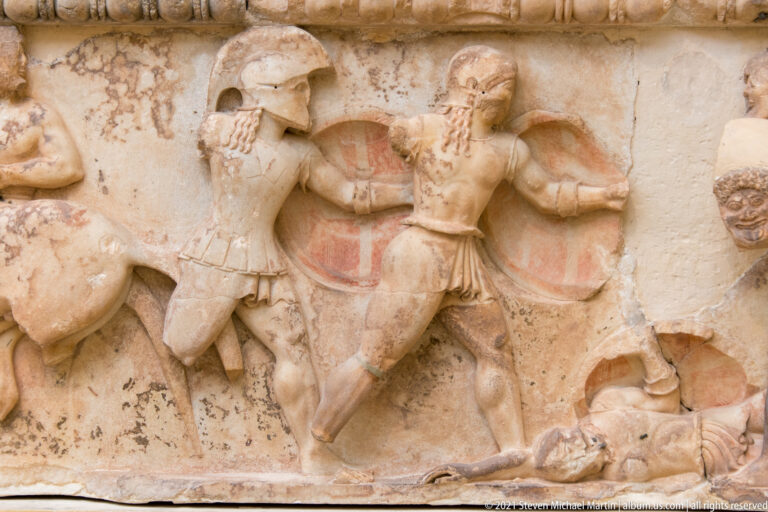 Archaic marble relief of two shield-bearing warriors fighting above a fallen figure, with traces of red pigment.